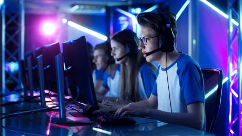Four high school students each wearing a headset with microphone and matching team tshirt sitting side by side in front of a monitor and keyboard while actively competing in an esports competition