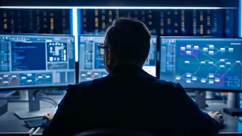 A close-up view of a person in a data center environment seated in front of three desktop monitors reviewing code and workflow displays