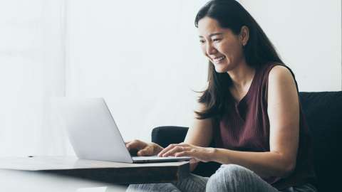 A woman sitting on a sofa smiles while typing on a laptop that rests on a coffee table