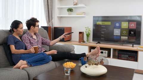 Two people sit next to each other on a sofa in a living room. One person uses a TV remote to navigate a streaming service selection menu displayed on a flat-screen TV