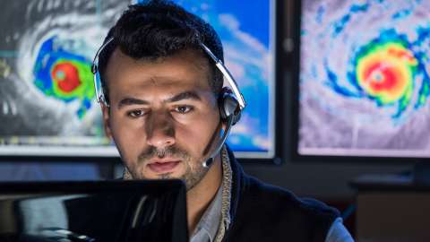 A close-up view of a seated developer wearing a headset and looking at a computer monitor. Two large monitors in the background display colorized weather satellite imagery