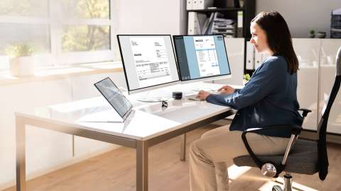 A small business owner sits at a desk in a modern, light-filled office reviewing invoices displayed on two side-by-side desktop monitors. A table displaying a spreadsheet is also on the desk