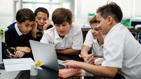 Five primary school students wearing school uniforms gather around an open laptop computer in a classroom environment