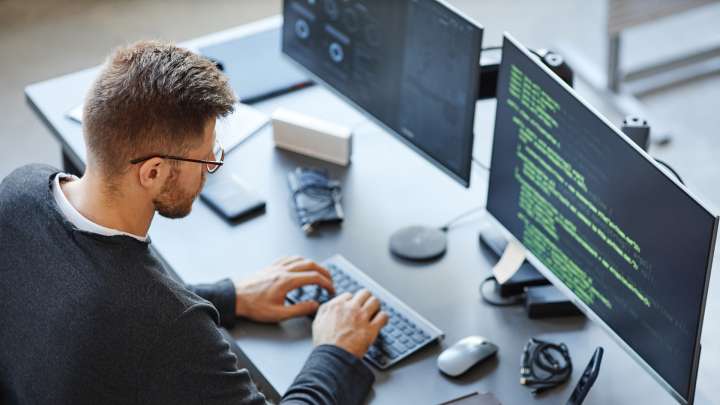 A cloud engineer sits at a desk typing on a keyboard. Code is shown on the desktop monitor