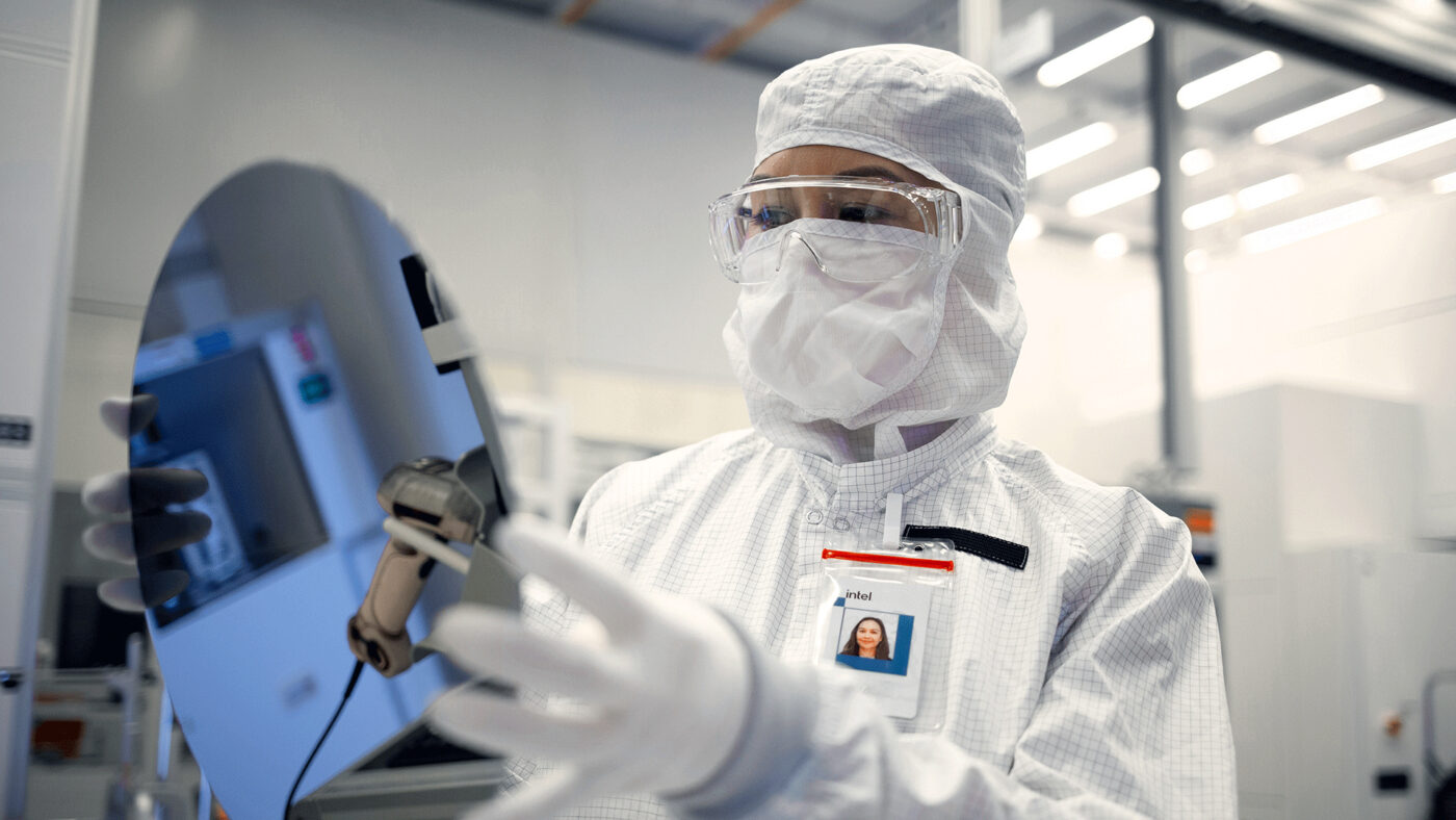 An Intel Foundry engineer in clean room gear holds a silicon waf