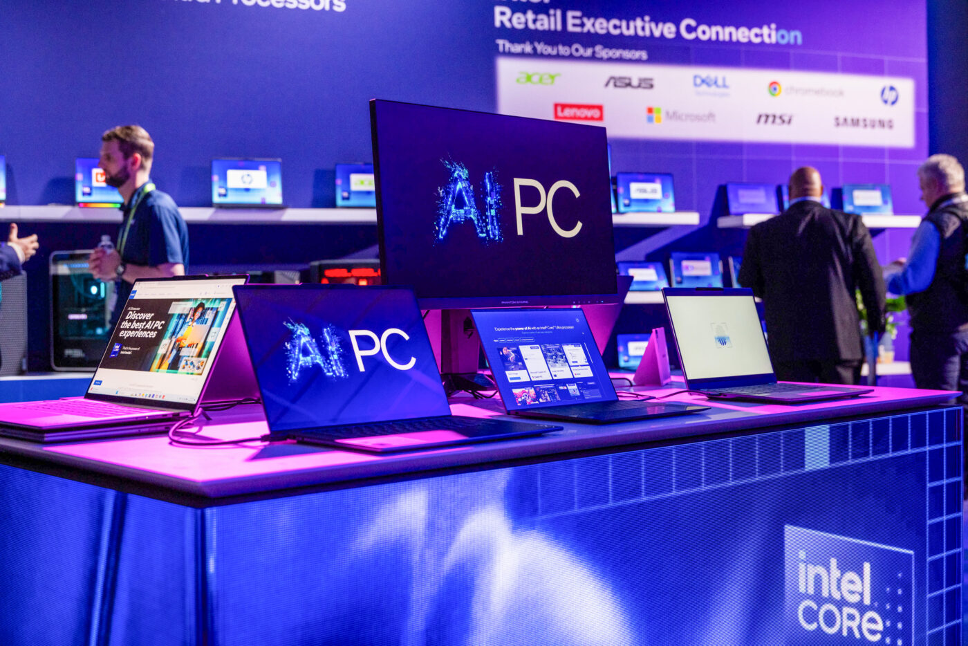 A display booth showcasing multiple laptops and monitors with AI PC graphics at a tech event. The setup is on a purple-lit table, with brand logos like Acer, ASUS, and Dell visible in the background. Attendees are in conversation nearby.