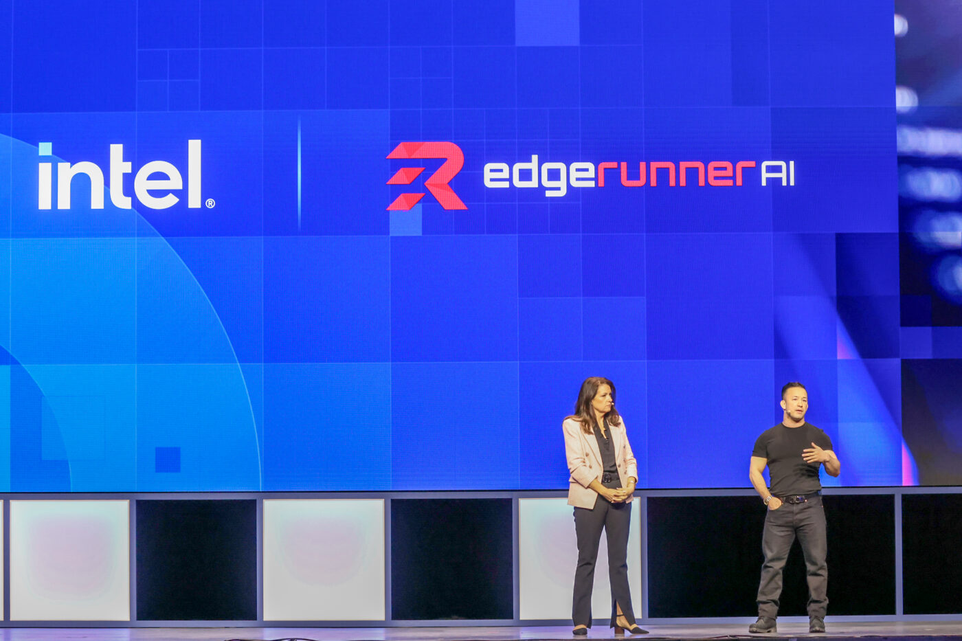 Two speakers stand on stage in front of a large screen displaying logos for Intel and EdgeRunner AI. The screen is blue with geometric patterns. The woman on the left is wearing a blazer, and the man on the right is in black attire.