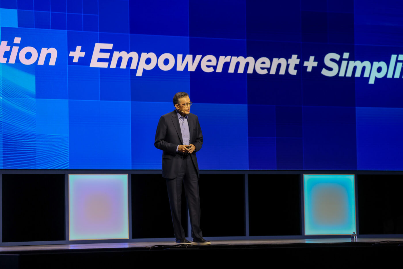 A person in a dark suit stands on a stage in front of a large blue screen with the words Innovation + Empowerment + Simplification. The stage is lit with soft, multicolored lighting from below.