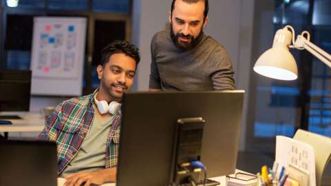 Two coworkers in a modern, open workspace collaboratively review information displayed on a desktop monitor. One person is seated and smiling while typing on a keyboard while the other stands and points at something on the screen