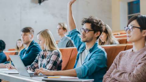 Close up view of university students seated in a lecture hall. One student has one arm raised while the other hand rests on an open laptop’s keyboard
