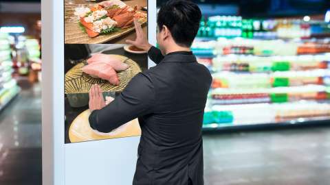 Person standing at digital kiosk in supermarket using hand gestures to enlarge photo of available products