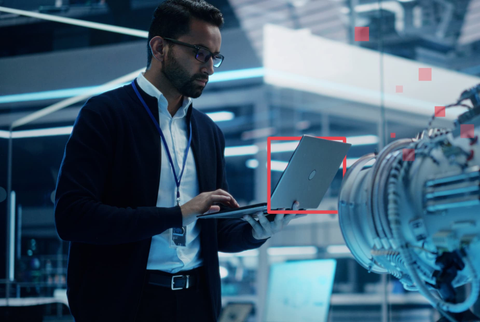 A man stands working on a laptop in a glass office and standing near a large microscope; stylistic red squares highlight the technology.